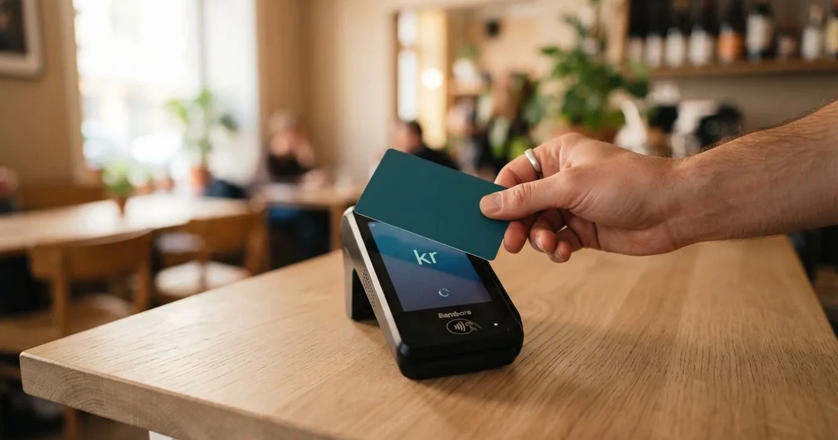 En hand håller ett betalkort nära en kortterminal för en kontaktlös betalning, fotograferat i närbild med varm belysning.
